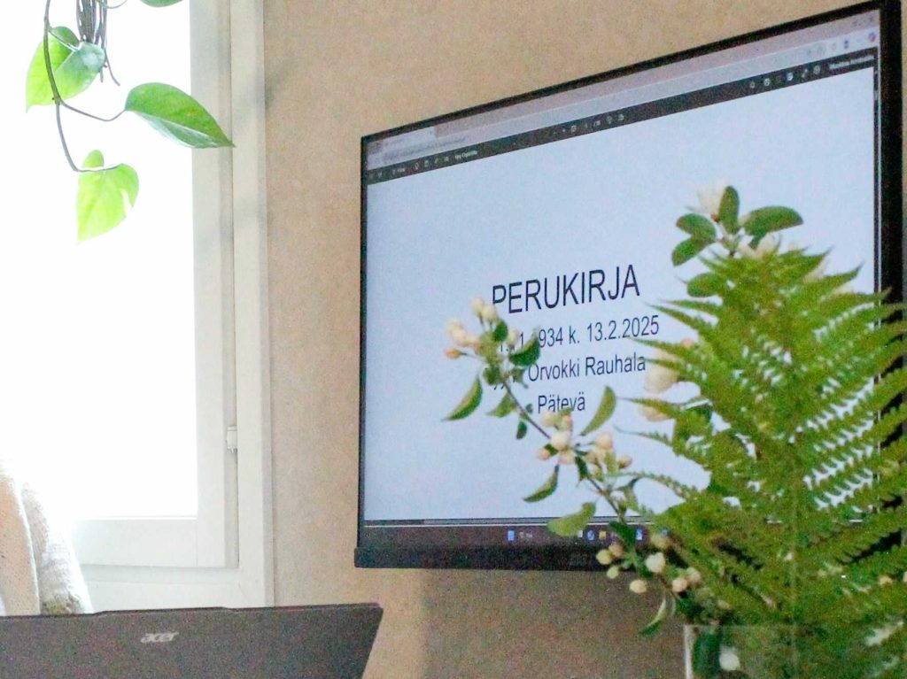 Seinälle kiinnitetyllä näytöllä on kuvitteellisen perukirjan kansilehti. Vasemmassa alakulmassa näkyy hieman kannettavan tietokoneen näytön yläreunaa, oikeassa reunassa omenankukan ja saniaisen lehti pehmentävät kauniisti näytöllä näkyvää näkymää.