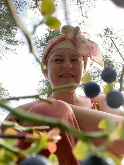 Mustikanvarpujen ja mustikoiden alta otettu kuva suoraan ylöspäin. Kuvassa Marja on polvistuneena mustikoiden yläpuolelle, taustalla näkyy kauniin kirkas taivas.